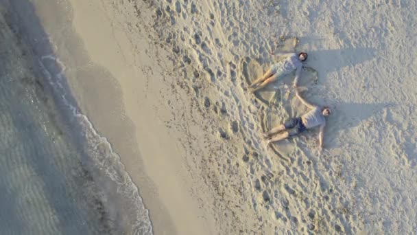 Um casal fazendo anjos na areia ao pôr do sol, aéreo — Vídeo de Stock