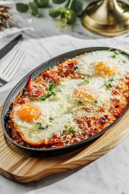 Geleneksel Shakshuka yumurta, domates ve maydanozlu hafif mermer arka planda demir bir tavada. Sağlıklı deniz ürünleri, sert ışık, restoran dekorasyonu, yakın çekim.