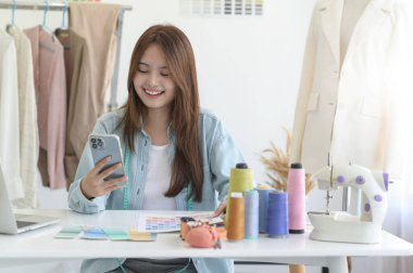 Bir tasarımcının atölyesinde dizayn ekipmanı, dizüstü bilgisayarı ve akıllı telefonu olan genç bir kadın terzi..