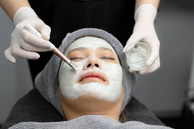 Güzellik uzmanı, güzellik kliniğinde krem maskeli genç bir kadın için krem maske kullanıyor..