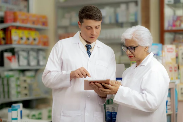 Young caucasian pharmacist and indian female pharmacist discussing, looking down at tablet in hand, drug consultant, front view.