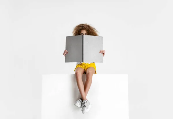 Portret małej ślicznej kręconej dziewczynki w luźnych ciuchach siedzącej na dużym pudełku i czytającej książkę izolowaną na białym tle studia. — Zdjęcie stockowe