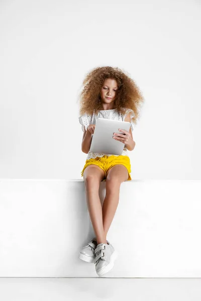 Una pequeña chica rizada linda en ropa casual sentada en una caja grande con tableta digital, gadget aislado en fondo de estudio blanco. — Foto de Stock