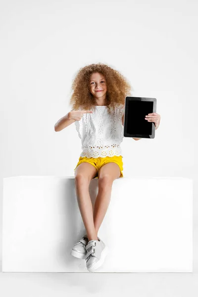 Una pequeña chica rizada linda en ropa casual sentada en una caja grande con tableta digital, gadget aislado en fondo de estudio blanco. —  Fotos de Stock