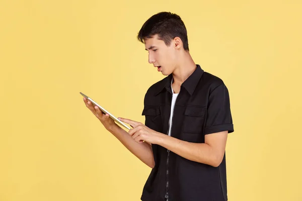 Portrait de jeune garçon, adolescent ou étudiant en tenue décontractée, chemise noire et jean bleu à l'aide d'une tablette isolée sur fond de studio jaune. — Photo