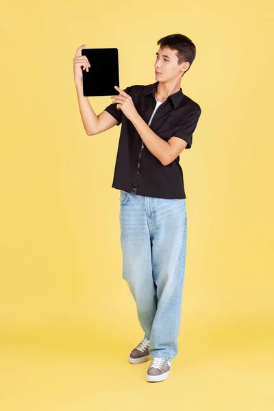 Sarı stüdyo arka planında izole edilmiş tablet kullanan genç, genç ya da öğrenci portresi, siyah gömlek ve kot pantolon.. — Stok fotoğraf