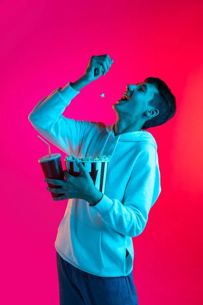 Jeden mladý chlapec, student, teen v neformálním oblečení drží obrovský kbelík popcornu izolované na fialovém pozadí v neonovém světle. — Stock fotografie
