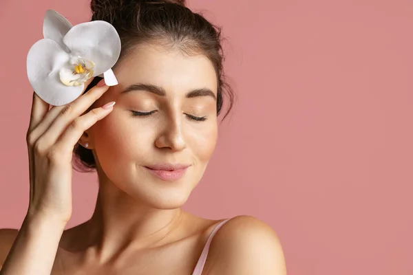 Close-up portret van een mooie glimlachende vrouw met witte bloem geïsoleerd over roze studio achtergrond. Natuurlijke schoonheid en esthetische kosmetologie concept. — Stockfoto