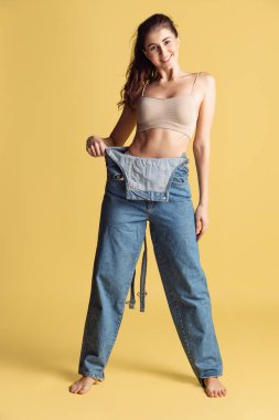 Sarı stüdyo arka planında izole edilmiş, mavi kot pantolonlu, genç ve zayıf bir kadının portresi. Kilo kaybı konsepti.