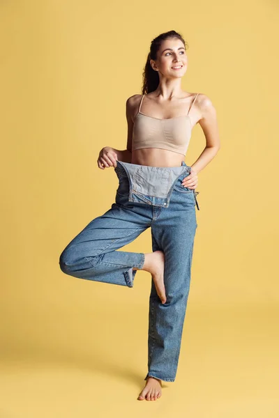 Portret van jonge mooie slanke vrouw in blauwe jeans jumpersuit poserend geïsoleerd over gele studio achtergrond. Gewichtsverlies concept. — Stockfoto