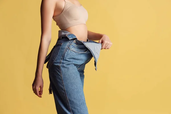 Ritratto di giovane bella donna magra in tuta blu jeans posa isolata su sfondo giallo studio. Perdita di peso concetto. — Foto Stock