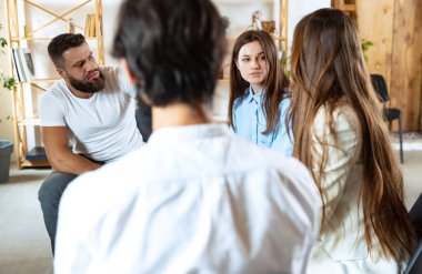 Genç çok ırklı insanlar, kadın ve erkekler birlikte oturuyor, konuşuyor, psikolojik destek grubunda, kapalı alanda iletişim kuruyor..
