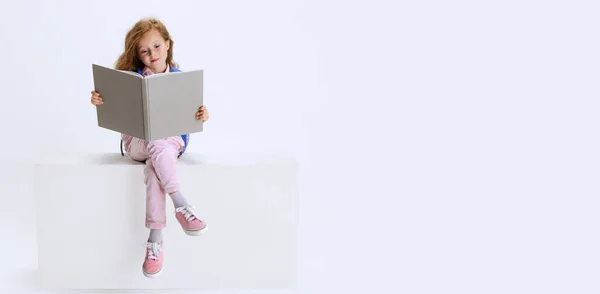 Jeden mały cute girl w casual ubrania siedzi na dużym pudełku i czytania książki izolowane na białym tle studio. — Zdjęcie stockowe