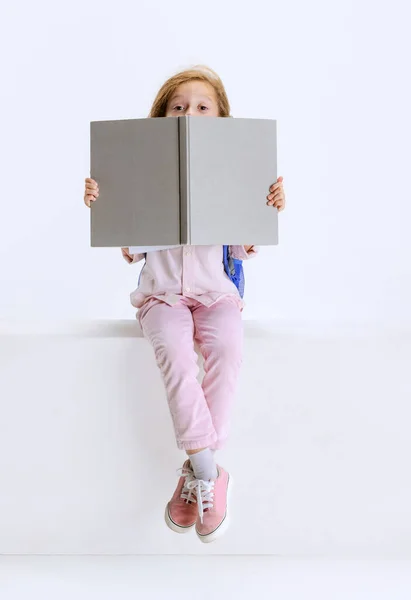 Una niña linda con ropa casual sentada en una caja grande y libro de lectura aislado en el fondo blanco del estudio. — Foto de Stock
