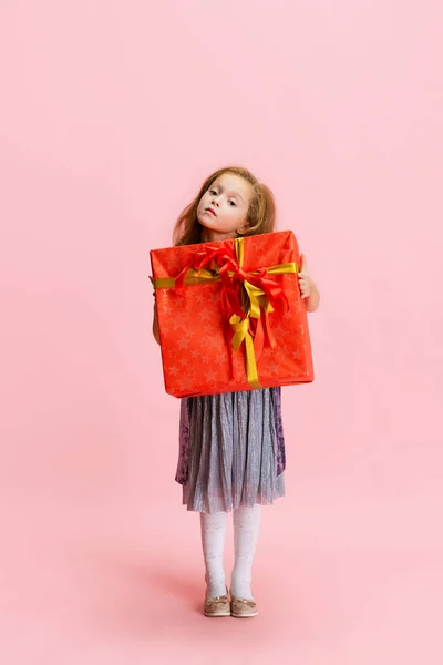 Um feliz bonito caucasiano menina weraing vestido de férias de pé com enorme presente, caixa de presente isolado no fundo do estúdio rosa. — Fotografia de Stock