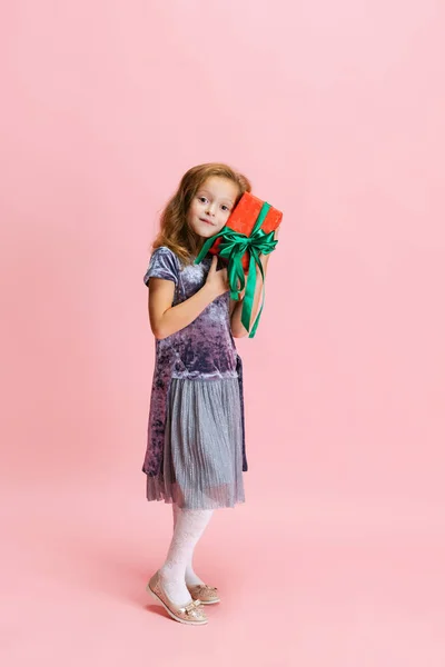 One happy cute caucasian little girl weraing holiday dress standing with huge gift, present box isolated on pink studio background. — Stock Photo, Image