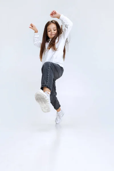 Full-length portrait of little cute girl, kid wearing hoodie with jeans steps, jumps isolated over white studio background. Stock Image