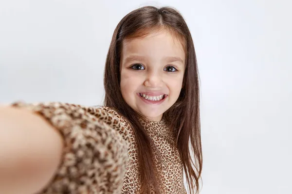 Vista frontal de la cámara de la pequeña niña linda sonriente preescolar hermosa tomando selfie aislado sobre fondo de estudio blanco. —  Fotos de Stock