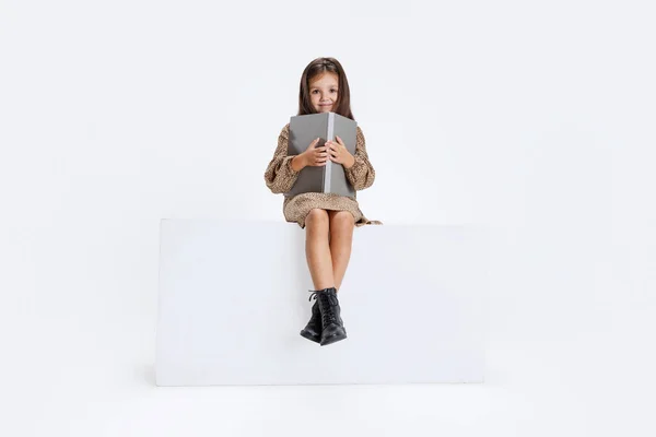 Portret małej ślicznej kręconej dziewczynki w luźnej sukience siedzącej na dużym pudełku i czytającej książkę izolowaną na białym tle studia. — Zdjęcie stockowe