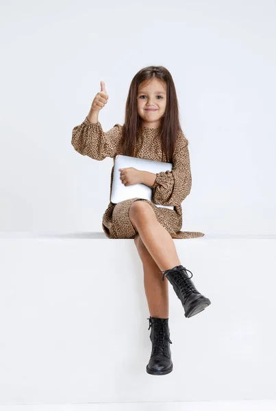 Une petite jolie fille mignonne, enfant en robe décontractée assis sur une grande boîte avec tablette numérique isolé sur fond de studio blanc. — Photo