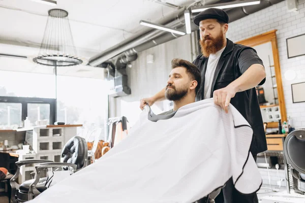 Dos hombres guapos en la barbería. Elegante peluquero barbudo rojo y cliente durante el proceso de corte de pelo. Concepto de belleza, salud y cosmética masculina. — Foto de Stock