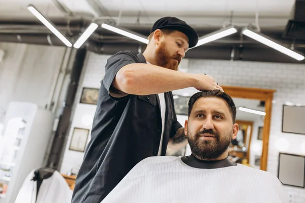 Två stiliga män på frisersalongen. Snygg rödskäggig frisör och klient under klippningsprocessen. Skönhet, sjukvård och manlig kosmetika koncept. — Stockfoto