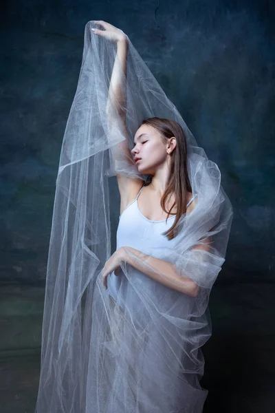 Portrait of young beautiful girl, ballerina posing with white transparent cloth isolated on dark vintage studio background. — Stock Photo, Image