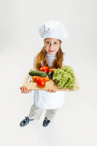 Vista de alto ângulo da menina fofa em uniforme cozinheiro branco e enorme chapéu de chefs segurando bandeja com legumes saudáveis isolados no fundo do estúdio branco. — Fotografia de Stock