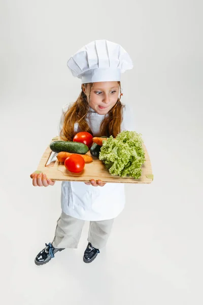 High angle view of little cute girl in white cook uniform and huge chefs hat holding tray with healthy vegetables isolated on white studio background. — Stock Photo, Image