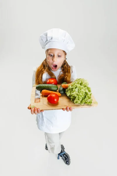 Vista de alto ângulo da menina fofa em uniforme cozinheiro branco e enorme chapéu de chefs segurando bandeja com legumes saudáveis isolados no fundo do estúdio branco. — Fotografia de Stock