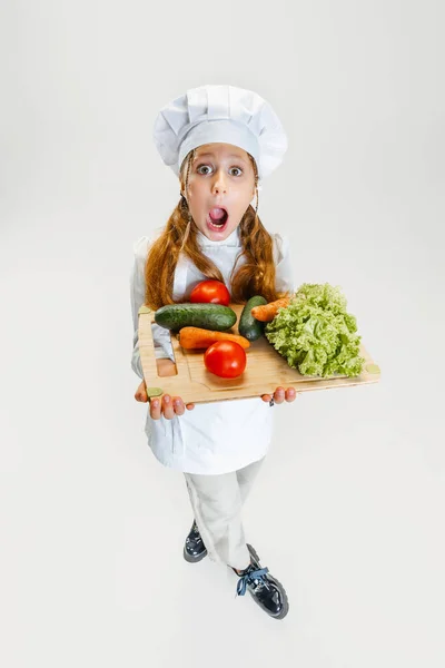 High angle view of little cute girl in white cook uniform and huge chefs hat holding tray with healthy vegetables isolated on white studio background. — Stock Photo, Image