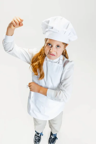 Hochwinkelblick auf kleines nettes Mädchen in weißer Koch-Uniform und riesigen Kochmütze posiert isoliert auf weißem Studiohintergrund. — Stockfoto
