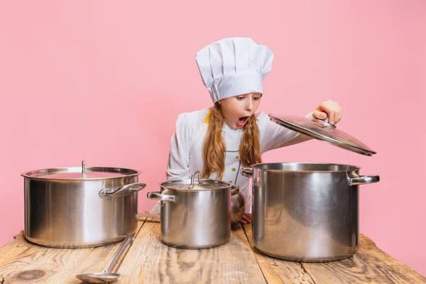 백인 요리사 복장의 어린 소녀와 거대 한 요리사 모자를 쓰고 핑크 스튜디오 배경에 커다란 냄비가 따로 있는 아이들 부엌에서. — 스톡 사진