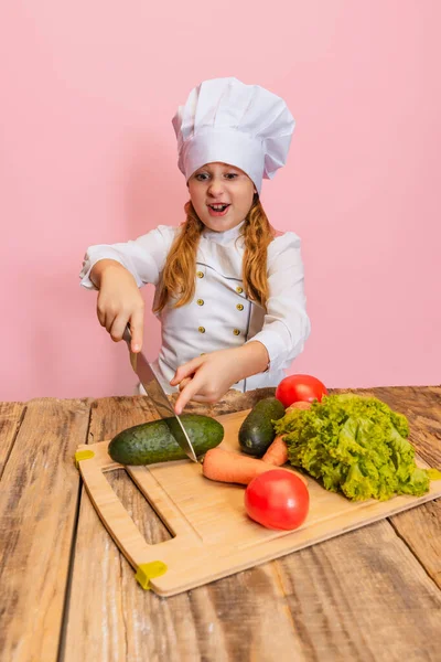 Young little girl in white cook uniform and huge chefs hat cutting fresh vegetables isolated on pink studio background. — Stock Photo, Image
