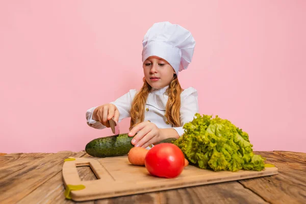 Young little girl in white cook uniform and huge chefs hat cutting fresh vegetables isolated on pink studio background. — Stock Photo, Image