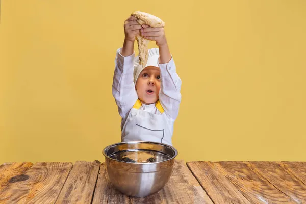 Lindo niño sonriente en cocinero blanco, chaqueta de chef y sombrero amasa la masa aislada en el fondo del estudio amarillo. — Foto de Stock