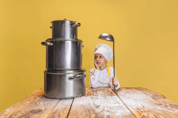 Primer plano lindo niño en uniforme de cocinero blanco y enorme sombrero de chefs en la cocina de los niños con grandes sartenes aislados en el fondo del estudio amarillo. — Foto de Stock