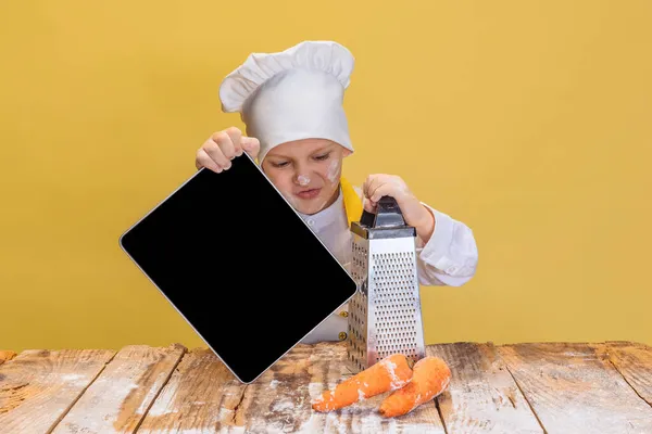 Close-up schattig jongetje in wit kokkin uniform en enorme chef-koks hoed op kinderen keuken met behulp van tablet geïsoleerd op gele studio achtergrond. — Stockfoto