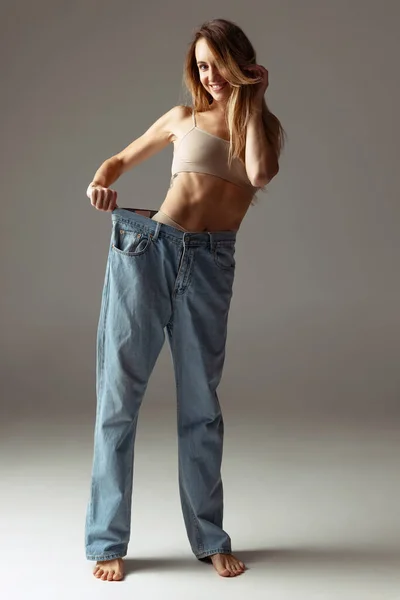 Retrato de joven hermosa mujer delgada en pantalones vaqueros azules jumpersuit posando aislado sobre fondo gris estudio. concepto de pérdida de peso. — Foto de Stock