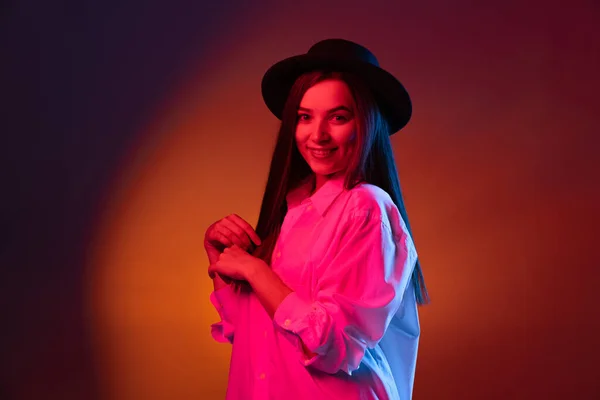 Stijlvol jong mooi meisje draagt zwarte hoed geïsoleerd op donkere studio achtergrond in neon licht. Concept van emoties, mode — Stockfoto