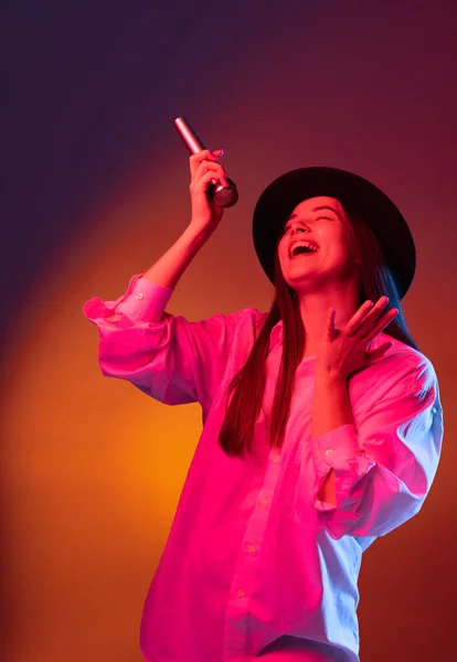 Een jong mooi meisje draagt zwarte hoed met behulp van telefoon geïsoleerd op donkere studio achtergrond in neon licht. Concept van emoties, mode — Stockfoto