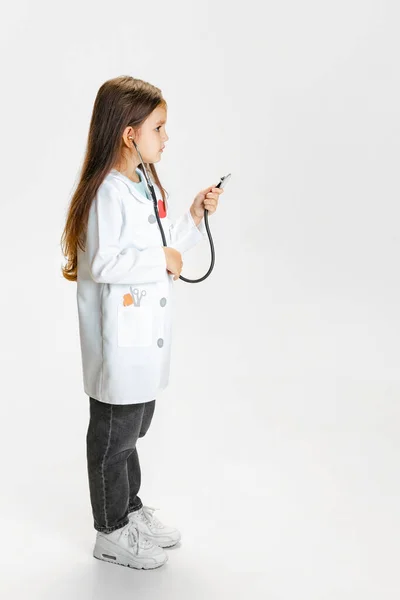 侧视图。漂亮的小女孩，穿着白色实验室外套的医生形象中的孩子，在白色工作室的背景下显得孤立无援 — 图库照片
