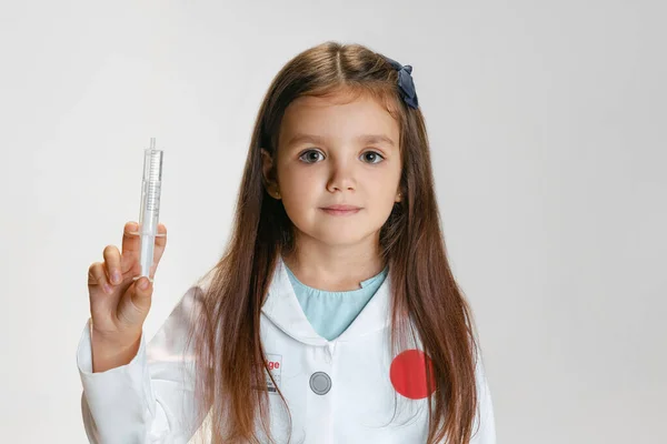 Портрет милой красивой девушки, ребенок в образе доктора в белом халате с игрушечным шприцем на белом фоне студии — стоковое фото
