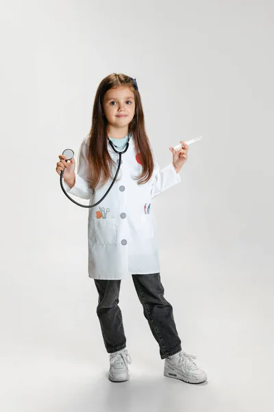 Portret van schattig mooi meisje, kind in beeld van arts dragen witte labjas poseren geïsoleerd op witte studio achtergrond — Stockfoto