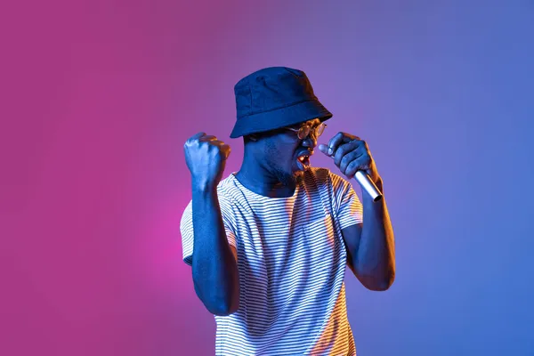 Студійний знімок емоційного африканського чоловіка з мікрофоном ізольовано над градієнтом фіолетового рожевого кольору студійного фону в неоновому фільтрі — стокове фото