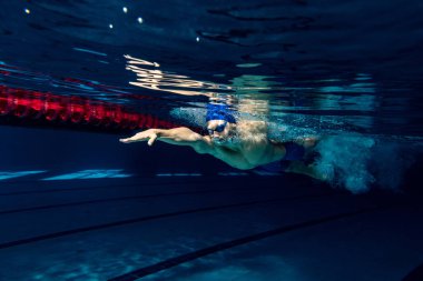 Yakın plan. Yüzme şapkalı ve gözlüklü genç bir erkek yüzücü havuz ve kapalı alanda antrenman yaparken hareket halinde ve hareket halinde. Su altı görüntüleme