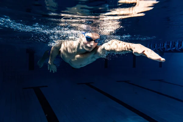 Närbild. Ung manlig simmare i badmössa och skyddsglasögon i rörelse och action under träning vid bassäng, inomhus. Undervattensavbildning — Stockfoto