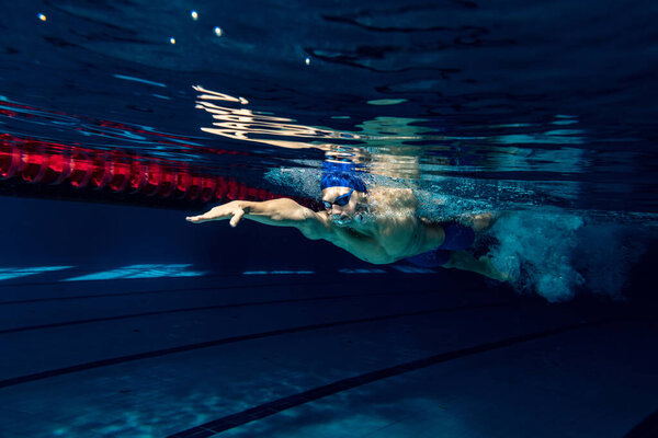 Close-up. Young male swimmer in swimming cap and goggles in motion and action during training at pool, indoors. Underwater imaging