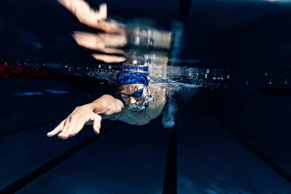 Närbild. Ung manlig simmare i badmössa och skyddsglasögon i rörelse och action under träning vid bassäng, inomhus. Undervattensavbildning — Stockfoto