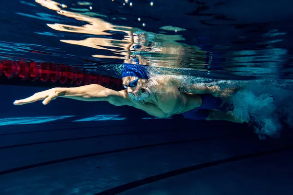 Nadador masculino profesional en gorra de natación y gafas en movimiento y acción durante el entrenamiento en la piscina, en interiores. Estilo de vida saludable, poder, energía, concepto de movimiento deportivo — Foto de Stock